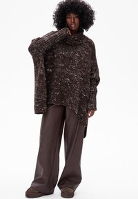 Kobieta z kręconymi włosami, ubrana w brązowy, oversize'owy sweter z dzianiny, szerokie brązowe skórzane spodnie i dopasowane brązowe buty, stojąca na białym tle.