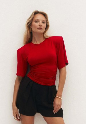 Kobieta z długimi blond włosami, w czerwonym marszczonym topie z krótkim rękawem i czarnych szortach z wysokim stanem, stoi przy białej ścianie.