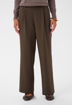 Femme portant un pantalon large vert olive, un pull marron, des chaussures plates foncées, les mains détendues le long du corps sur un fond uni.