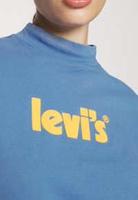 Modrá bavlněná mikina s rolákem, která se vyznačuje výrazným žlutým logem "levi's" zaobleného písma, hladkým povrchem a ribovaným límcem.