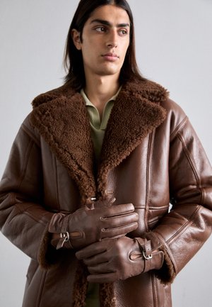 Brun läderjacka i shearling med en tjock, texturerad krage och matchande bruna handskar, som har en spänne-detajl vid handleden.