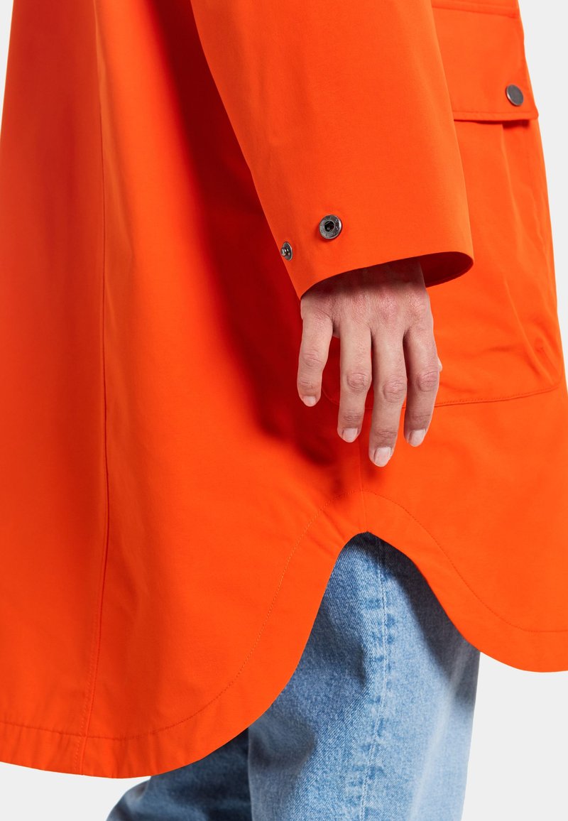 Leuchtender orangefarbener Mantel mit abgerundetem Saum, Druckknopfmanschetten und strukturiertem Stoff. Hand liegt an der Seite. Über leichter Denim getragen.
