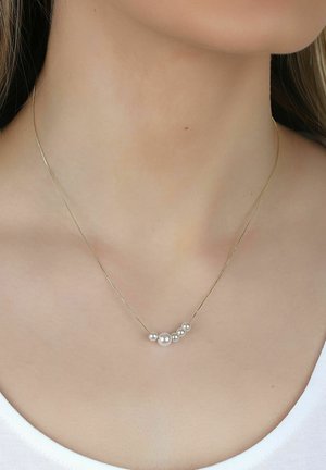 Zarte goldene Halskette mit einer gebogenen Reihe von sieben weißen Perlen in verschiedenen Größen am Hals einer Frau mit weißem Top.
