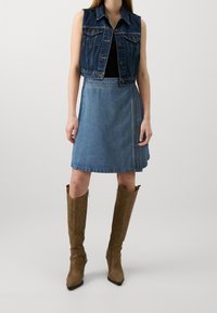 Jeansväst med ärmlös design, ljus tvättad A-linje kjol och tan knähöga stövlar. Har knappstängningar och två bröstfickor.