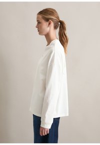 Weißer Langarmpullover mit lockerer Passform, Rundhalsausschnitt und dezentem Logodetail auf der Brust. Kombiniert mit blauen Jeans.