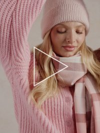 Ljusrosa stickad tröja med ribbad textur och knappar, kombinerad med en rutig halsduk i rosa toner och en matchande rosa mössa.