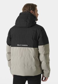Gevoerde jas met een zwarte bovenkant en een beige onderkant, voorzien van een capuchon en een gestructureerde afwerking, gelabeld met "HELLY HANSEN" op de achterkant.