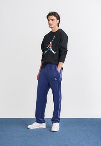 Μαύρη μπλούζα με χρωματιστό λογότυπο, συνδυασμένη με μπλε φόρμες με λευκή ρίγα στο πλάι και λευκά αθλητικά παπούτσια. Απλή, casual αθλητική ένδυση.