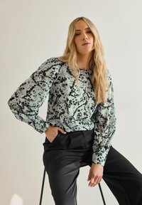 Blusa azzurra con motivi astratti neri, maniche lunghe con polsini arricciati, abbinata a pantaloni neri a gamba larga. Design semplice.