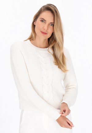 Vrouw met lang blond haar in een witte, gestructureerde trui, die de mouw aanpast tegen een effen witte achtergrond.