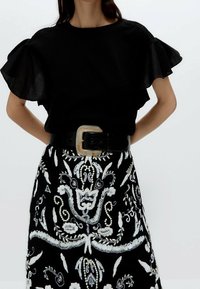 Haut noir à manches volantes, associé à une jupe brodée noir et blanc ornée de motifs floraux complexes et d'une boucle de ceinture décorative.