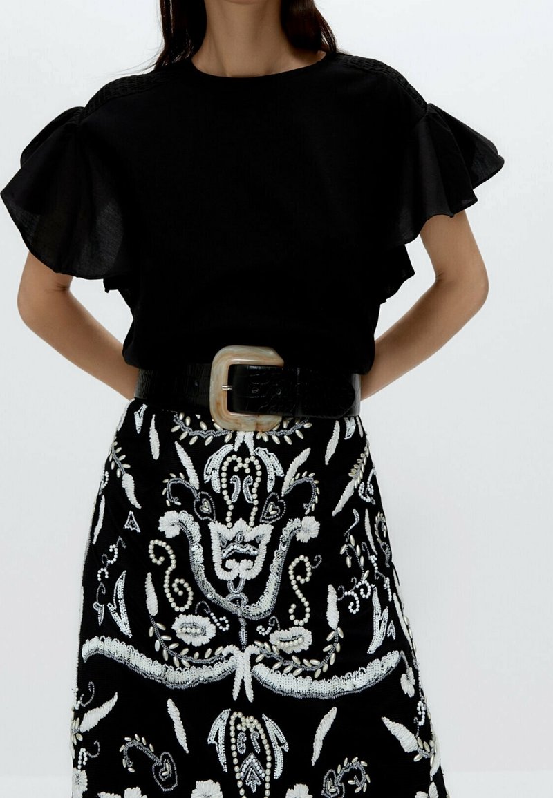 Haut noir à manches volantes, associé à une jupe brodée noir et blanc ornée de motifs floraux complexes et d'une boucle de ceinture décorative.