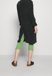 Lång svart kappa med slits i ryggen, i kombination med gröna korta byxor och svarta sandaler. Tyget ser lätt och slätt ut.