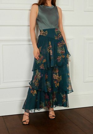 Falda maxi acampanada de flores en verde azulado oscuro con volantes, combinada con una blusa de seda gris. Presenta intrincados patrones florales y un diseño en capas.