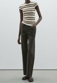 Randig stickad topp i kräm, brun och beige, kombinerad med mörka, avslappnade byxor. Modellen bär bruna sandaler. Släta texturer är tydliga.