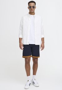 Hvit skjorte over en enkel hvit t-skjorte, kombinert med marineblå shorts med brun oppbrett. Svarte solbriller og hvite joggesko.