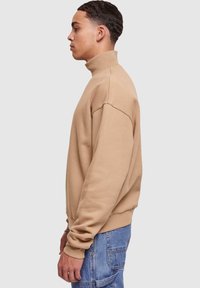 Felpa beige con collo alto, spalle scese, polsini a coste e vestibilità rilassata, abbinata a jeans in denim blu.