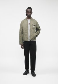 Vyras dėvi šviesiai alyvinę siuvinėtą striukę ant pilkos grafikos marškinėlių, juodus kelnes ir juodus batus, stovintis priešais paprastą baltą foną.