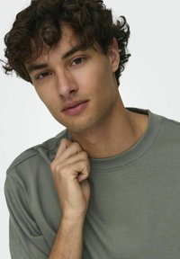 Chemise à manches courtes vert olive en tissu doux avec un col rond et des détails de couture discrets, portée par une personne aux cheveux bouclés.