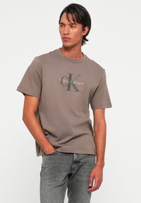 Mlad čovjek s kratkom tamnom kosom koji nosi smeđu majicu Calvin Klein i sive traperice, gleda u stranu s jednom rukom u džepu.