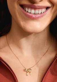 Gouden ketting met een strikpendant met gebogen randen, weergegeven op een blote hals tegen een kastanjebruine top. Gladde metalen oppervlakte.