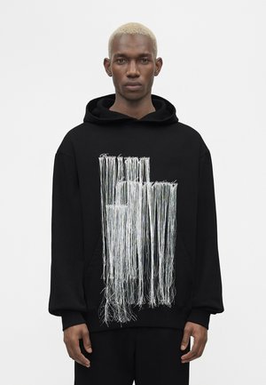 Czarna bluza z kapturem noszona przez stojącą osobę z krótkimi blond włosami, z długimi białymi frędzlami zwisającymi nad przednim napisem.