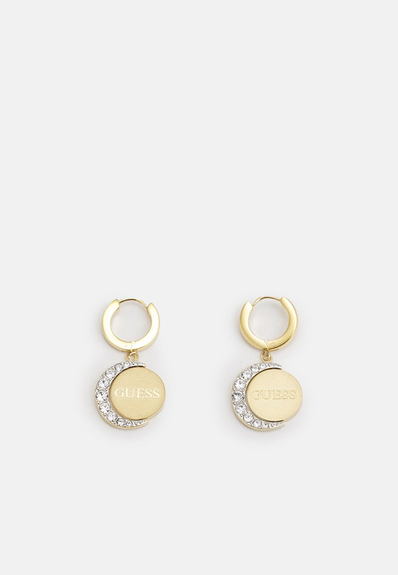 Des boucles d'oreilles en métal doré en forme de créole présentent des disques ronds avec une gravure "GUESS" et un design en croissant orné de strass transparents.