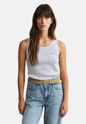 Frau mit langen braunen Haaren, die ein hellblaues ärmelloses Top mit Zopfmuster, einen gewebten beigen Gürtel und eine hoch taillierte blaue Jeans vor weißem Hintergrund trägt.