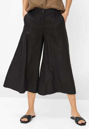 Kvinde iført sorte cropped bukser med brede ben, frontknapper og sidelommer, kombineret med sorte sandaler med krydsremme.