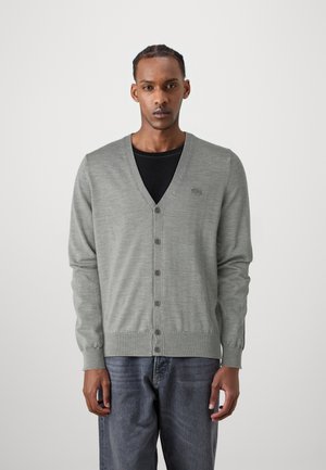 Cárdigan de punto gris con cuello en V, seis botones y puños acanalados. Presenta un pequeño logo en el lado izquierdo del pecho. Llevado sobre una camisa negra.