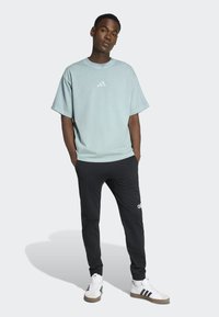 Άνδρας που φοράει ανοιχτό μπλε oversized t-shirt, μαύρο παντελόνι φόρμας Adidas και λευκά παπούτσια Adidas, στέκεται με τα χέρια στις τσέπες.
