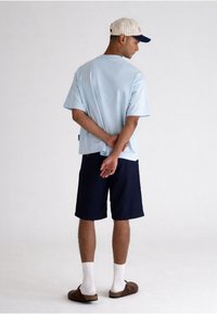 Homme debout, les mains derrière le dos, portant un t-shirt bleu clair, un short bleu marine, des chaussettes blanches, des sandales marron et une casquette de baseball rayée.