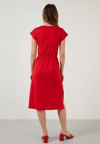 Rotes Baumwoll-Midikleid mit kurzen Ärmeln, elastischem Bund, seitlichen Schlitzen und glatter Textur. Kombiniert mit roten Blockabsatzsandalen.