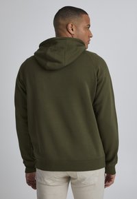 Sudadera verde oliva con capucha ajustable, puños y dobladillo acanalados, y una textura suave. Vista desde la parte trasera, combinada con pantalones de color claro.