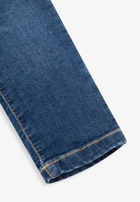 Jeans de mezclilla azul oscuro con corte recto. El dobladillo presenta una costura de color naranja claro en contraste a lo largo del borde para añadir un detalle adicional.