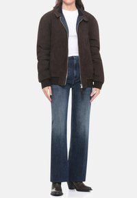 Giacca bomber in camoscio marrone con polsini e collo a costine, abbinata a jeans a gamba larga in denim blu a vita alta e stivali scuri con tacco.