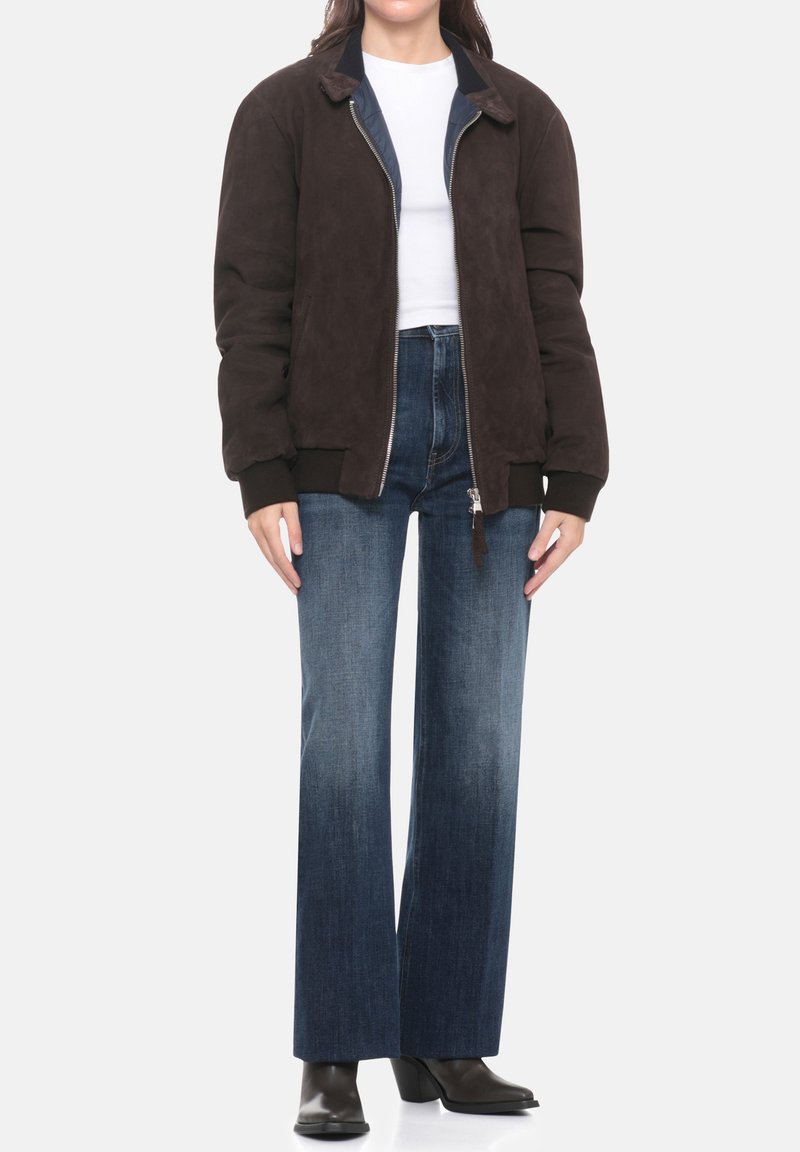 Giacca bomber in camoscio marrone con polsini e collo a costine, abbinata a jeans a gamba larga in denim blu a vita alta e stivali scuri con tacco.