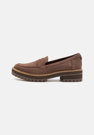 Mocasín de cuero marrón con suela de tread grueso y tacón bajo, que presenta un detalle de correa en la parte superior y costuras visibles.