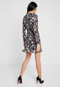 Vestido negro de flores con mangas largas de tul, que presenta patrones coloridos de margaritas, una falda acampanada y un dobladillo con volantes. Lleva consigo unos tacones altos negros.