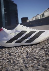 Weißer Adidas-Laufschuh mit drei schwarzen Streifen an der Seite, liegend auf strukturiertem Pflaster mit Gebäuden im Hintergrund.