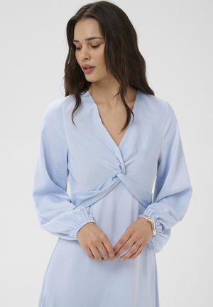 Femme aux longs cheveux foncés portant une robe bleu clair à manches longues avec un détail de nœud torsadé au niveau de la poitrine et des poignets élastiques, les mains jointes.