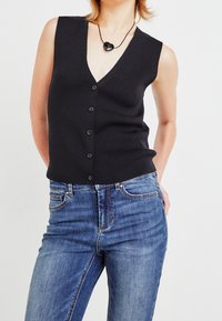 Cardigan sin mangas negro con escote en forma de V, cierre de botones en el delantero, textura acanalada, combinado con jeans azules. Collar negro en forma de corazón.
