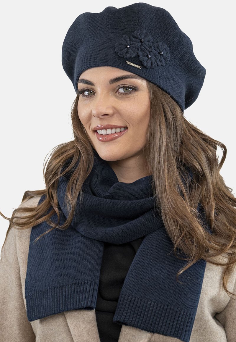 Berretto in maglia blu navy con dettagli floreali e un piccolo tag metallico; abbinato a una sciarpa blu navy coordinata con bordi a costine.