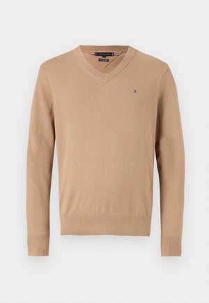 Beige v-hals genser laget av mykt stoff, med lange ermer og en liten logo på øvre venstre side.