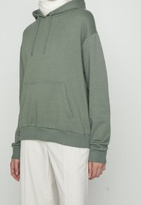 Persona vistiendo un sudadera verde salvia con bolsillo frontal y cordones, combinada con pantalones de pierna ancha en color blanco roto, frente a un fondo sencillo.