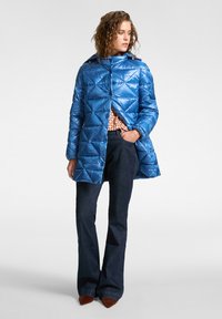 Vrouw gekleed in een blauwe gewatteerde jas, een patronen top, donkere flared jeans en bruine puntige schoenen, staande met één hand in haar zak en kijkend opzij.
