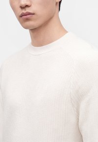Gros plan d'une personne portant un pull en tricot blanc texturé avec un col rond, montrant la partie inférieure de son visage et le haut de son torse.