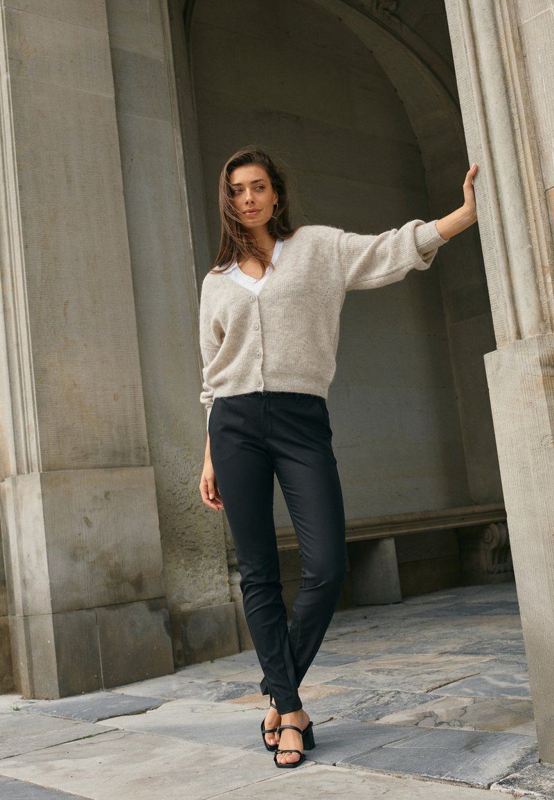 Hellbeige gestrickte Strickjacke mit Knöpfen, über einem weißen Top getragen, kombiniert mit schwarzen, figurbetonten Hosen und schwarzen, hohen Sandalen. Auffälliger Bogen als Hintergrund.