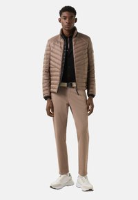 Polstret brun jakke med lynlåslukning foran, sort skjorte, beige bukser og hvide sneakers. Glat tekstur, tætsiddende design og kontrasterende farver.