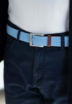 Ceinture tissée bleu clair avec boucle argentée portée avec un pantalon noir et une chemise blanche rentrée.